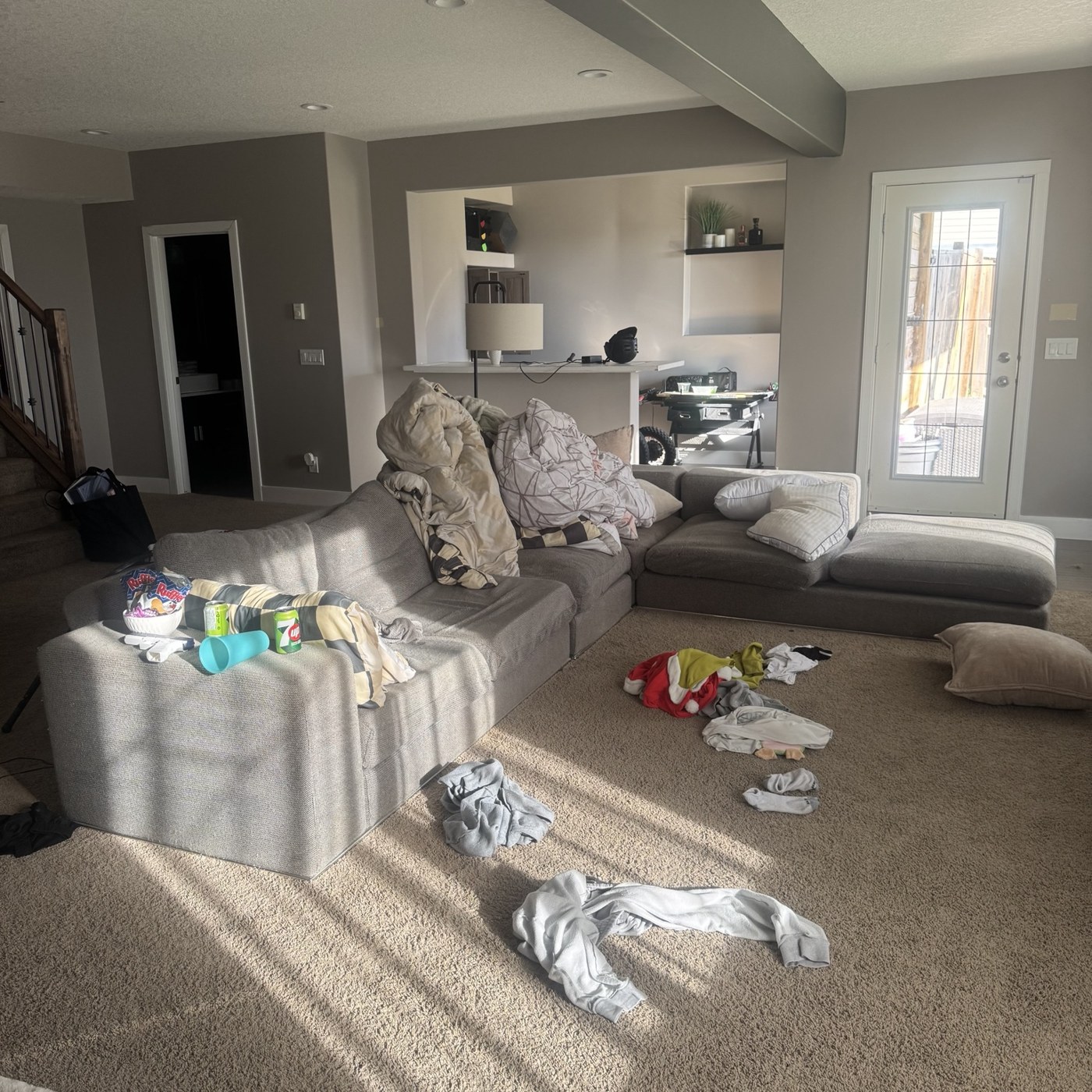 Living area before cleaning, with scattered clothing and items around a grey sectional sofa